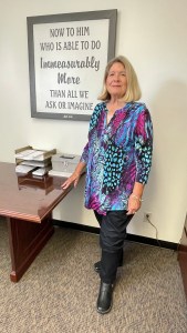 Carolyn Hamby stands near a wall hanging imprinted with words from the Bible.